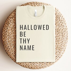 May include: A light beige t-shirt with the text "HALLOWED BE THY NAME" printed in black.