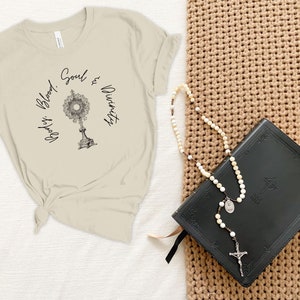 May include: A beige t-shirt with the text "Body, Blood, Soul & Divinity" and a graphic of a monstrance. A black leather-bound book and a white rosary are on a brown woven surface.