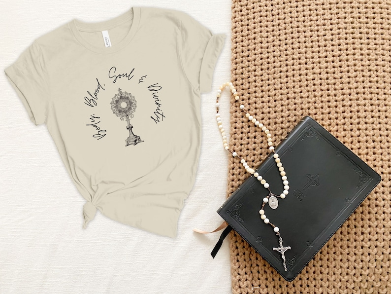 May include: A beige t-shirt with the text "Body, Blood, Soul & Divinity" and a graphic of a monstrance. A black leather-bound book and a white rosary are on a brown woven surface.