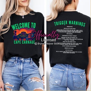 May include: Black t-shirt with the text "WELCOME TO CAPE CARNAGE" in green and a colorful graphic of a lighthouse and buildings. The back of the shirt has a list of "TRIGGER WARNINGS" in green.