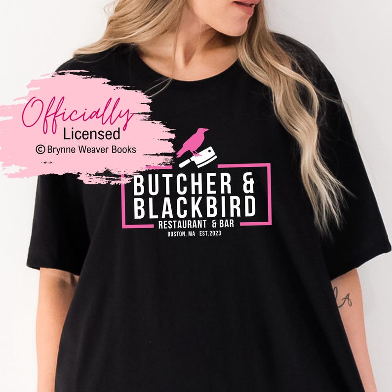 以下が含まれることがあります： 黒のTシャツに、ピンクのグラフィックデザインが施されています。デザインには「Butcher & Blackbird Restaurant & Bar Boston, MA Est. 2023」の文字と、ピンクの鳥がピンクの肉屋のナイフにとまっている様子が描かれています。