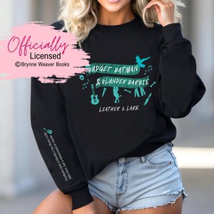 Op de afbeelding: Zwart sweatshirt met een turquoise grafische print met de tekst "Budget Batman & Blunder Barbie" met muzieknoten, een schedel, een microfoon, een gitaar, een bot, een jas en een mes. De tekst "Leather & Lark" staat onder de print. De tekst "Officially Licensed" staat in roze in de linkerbovenhoek van de afbeelding.