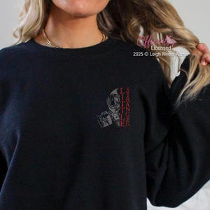 Op de afbeelding: Zwarte sweatshirt met een gasmasker ontwerp en de tekst "LITTLE STRANGER" in het rood. De afbeelding staat op de linkerbovenborst. De sweatshirt is effen zwart.