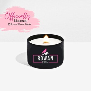 May include: A black candle with a pink logo that says "ROWAN Butcher & Blackbird" and a pink bird with a knife. The candle is lit and the flame is visible.