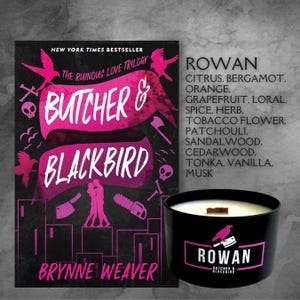 May include: A black candle with a wooden lid and a pink and black label that says "Rowan Butcher & Blackbird". The candle is sitting on a table in front of a book cover with the title "Butcher & Blackbird" and the author's name "Brynne Weaver".