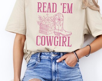 Peut inclure: T-shirt beige avec l'inscription rose "READ 'EM COWGIRL". Le motif comprend une pile de livres, une botte de cow-boy et des éléments floraux. Le t-shirt est porté avec un jean bleu.