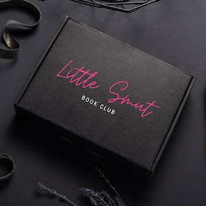 May include: A black cardboard box with the text "Little Smut Book Club" in pink script. The box is closed and positioned on a dark surface, with decorative elements in the background. The overall aesthetic is modern and minimalist.