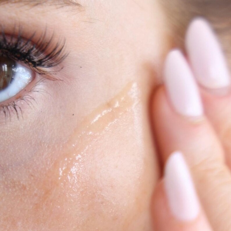 May include: Close-up of a person's face with a clear, shiny serum or moisturizer applied to the cheek. The serum is being rubbed in with a finger.