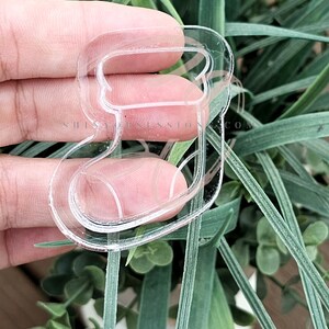 May include: Clear acrylic ornament shaped like a baby's foot, set against green leaves. The ornament features a curved top and a rounded foot design. The text "SHINYOBSESSION.COM" is printed on the ornament.