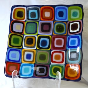May include: A square glass dish featuring a grid of multicolored nested square patterns. Each square contains concentric rings of various colors including blue, green, orange, red, purple, and yellow, set against a dark blue background.
