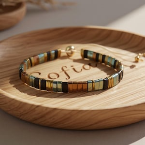 May include: A beaded bracelet with a mix of black, gold, and teal square beads. The bracelet is displayed on a wooden tray with the name "Sofia" engraved on it. The bracelet has a gold-colored clasp.