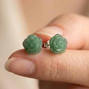 May include: A pair of stud earrings featuring carved light green stone roses. Each rose-shaped gemstone is mounted on a small silver-toned metal post. The earrings display a smooth, polished surface with intricate petal details in a soft green hue.