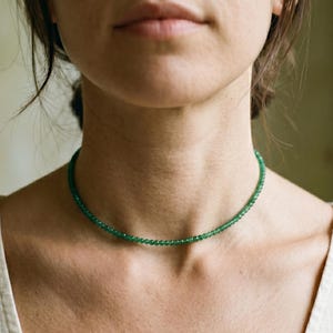 May include: A delicate, emerald green beaded necklace. The necklace is a choker style, resting closely around the neck. The beads are small and evenly spaced, creating a continuous line of color. The necklace is worn against a neutral background.