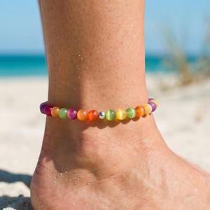 May include: Ankle bracelet featuring colorful, round beads in shades of purple, green, orange, and yellow, strung together with a silver-colored metal clasp. The jewelry is worn on an ankle, with a beach and ocean background.