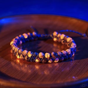 May include: A double-strand bracelet with amber-coloured beads and dark cord, presented on a wooden tray. The beads are tightly woven together. The background features a gradient of blue and brown, creating a warm contrast.