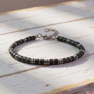 May include: A black beaded bracelet with silver-colored metal accents. The bracelet is made of small, cylindrical black beads interspersed with silver-toned metal beads. It has a silver-colored chain and clasp.
