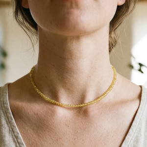 May include: A delicate, yellow beaded necklace. The necklace is composed of small, round beads, creating a subtle, elegant accessory. The necklace is worn around the neck, resting on the collarbone. The background is blurred, with soft lighting.