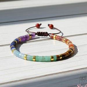 May include: A colorful beaded bracelet with a sliding knot closure. The bracelet features a variety of gemstones in a rainbow of colors, including purple, blue, green, and brown. Gold-colored spacers separate the beads. The bracelet is displayed on a white wooden surface.