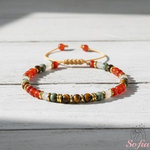 Op de afbeelding: Een kralenarmband met kleurrijke edelstenen, waaronder rode, bruine, groene en witte kralen. De armband heeft een schuifknoopsluiting en goudkleurige accenten. De armband ligt op een wit houten oppervlak.