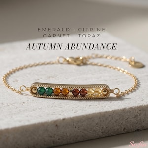 May include: A gold-tone chain bracelet featuring a central horizontal bar with a row of faceted beads in green, orange, dark red, and yellow. The text reads EMERALD CITRINE GARNET TOPAZ and AUTUMN ABUNDANCE. A small round tag is attached to the chain.