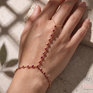 May include: A gold hand chain featuring a series of small, round, bezel-set red stones. The jewelry connects a delicate wrist bracelet to a single ring component, creating a continuous chain design that rests across the back of the hand.