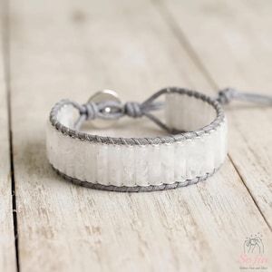 May include: A wrap bracelet featuring a single row of cylindrical white translucent stone beads secured with grey cord stitching. The bracelet includes a circular silver-toned metal button closure and a grey cord tie at the end for adjustment.