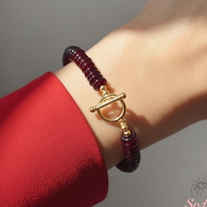 Op de afbeelding: Een bordeauxrode kralenarmband met een goudkleurige tuimelsluiting. De armband is gemaakt van kleine, ronde kralen en wordt om de pols gedragen. De sluiting heeft een cirkelvormig ontwerp met een staafsluiting.