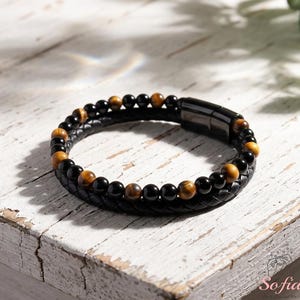 May include: A black leather bracelet with alternating black and tiger eye beads. The bracelet has a magnetic clasp and is displayed on a distressed white wooden surface. The bracelet is a piece of jewelry.