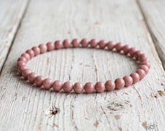 May include: A circular beaded bracelet composed of numerous small, smooth, dusty rose-colored spherical beads. The beads are uniform in size and shape, strung together to form a continuous loop. The bracelet is positioned flat, displaying its round form.