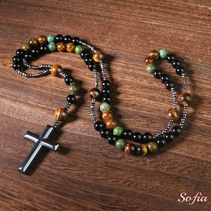 May include: A black, green, and brown beaded necklace with a silver cross pendant. The necklace is made of natural stones and is a stylish accessory for men.