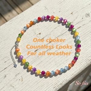 May include: A circular choker necklace featuring multicolored cat's eye beads in shades of red, orange, yellow, green, blue, and purple. The necklace includes a silver-toned metal chain extender with a small heart-shaped charm at the end.