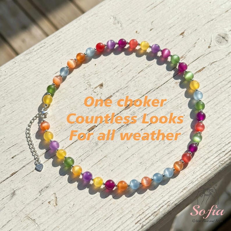 May include: A circular choker necklace featuring multicolored cat's eye beads in shades of red, orange, yellow, green, blue, and purple. The necklace includes a silver-toned metal chain extender with a small heart-shaped charm at the end.