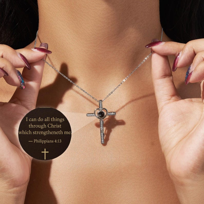 Puede incluir: Un collar con colgante de cruz plateada con un centro en forma de coraz&oacute;n y detalles brillantes. El collar se muestra sobre un fondo de tono de piel. Tambi&eacute;n es visible un gr&aacute;fico circular con texto de Filipenses 4:13.