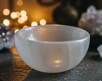 Può includere: Una ciotola di selenite bianca, un tipo di cristallo noto per la sua energia calmante, si trova su una superficie grigio scuro. La ciotola è liscia e lucida, con un morbido colore bianco latte.