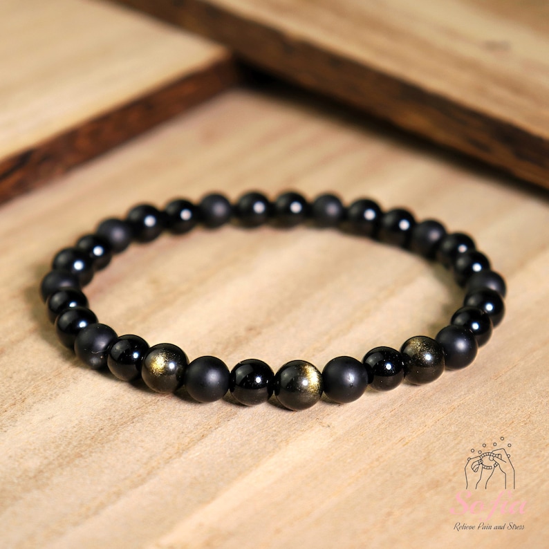 Peut inclure: Bracelet en perles noires avec des accents dor&eacute;s. Le bracelet est compos&eacute; de perles noires mates et de perles dor&eacute;es. Le bracelet est sur une surface en bois.