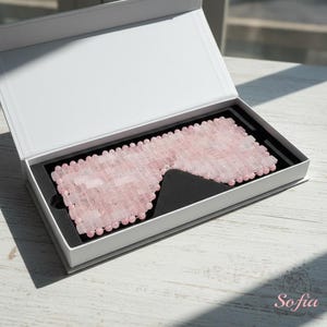 May include: A pink rose quartz eye mask is presented in an open white box with a black interior. The mask is composed of small, rectangular rose quartz stones, designed to cover the eyes. The box is on a white wooden surface.