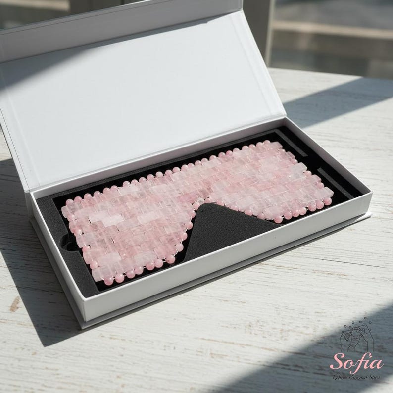May include: A pink rose quartz eye mask is presented in an open white box with a black interior. The mask is composed of small, rectangular rose quartz stones, designed to cover the eyes. The box is on a white wooden surface.