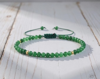 May include: Two green beaded bracelets. One bracelet is in focus, featuring small, round, green beads with gold accents. The other bracelet is out of focus, with a green cord and adjustable knot. The bracelets are on a white wooden surface.