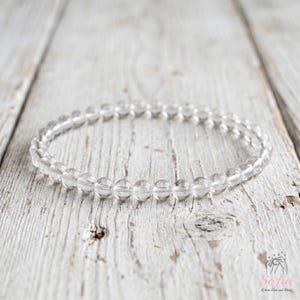 May include: A circular bracelet composed of small, clear, spherical beads strung together. The beads are translucent and uniform in size, creating a continuous loop. The bracelet is positioned horizontally, showcasing its simple, beaded construction.