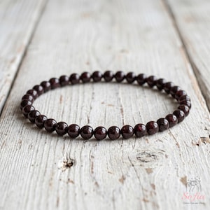 Peut inclure: Un bracelet circulaire composé de petites perles de grenat rouge foncé, lisses et enfilées ensemble. Les perles sont uniformes en taille et en forme, formant une boucle continue. Les pierres rouge profond ont un éclat subtil sur leurs surfaces.