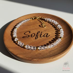 Könnte beinhalten: Ein zartes goldenes Kettenarmband mit klaren Quarzperlen und goldenen Akzenten. Das Armband liegt auf einer Holzoberfläche mit dem eingravierten Namen "Sofia".