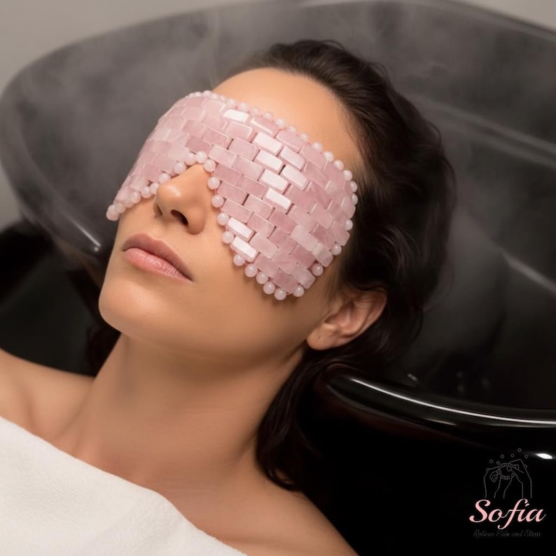 May include: A pink rose quartz eye mask with a woven design and rounded edges. The mask is made of small, rectangular stones and is being worn by a person. The mask is designed for relaxation and skincare.