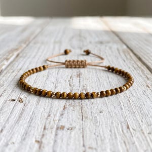 May include: A bracelet featuring small, round, golden-brown tiger eye beads interspersed with tiny metallic gold-tone spacer beads. The bracelet includes an adjustable sliding knot closure made from tan cord, finished with two small beads at the ends.