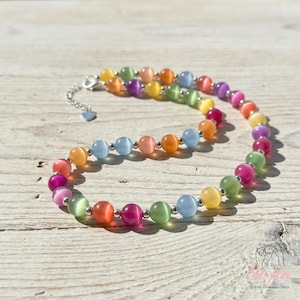 May include: A necklace featuring a strand of multicolored cat's eye beads in shades of pink, orange, yellow, green, and blue. The beads are separated by small silver-toned metal spacers, and the necklace includes a silver-toned metal chain extender.