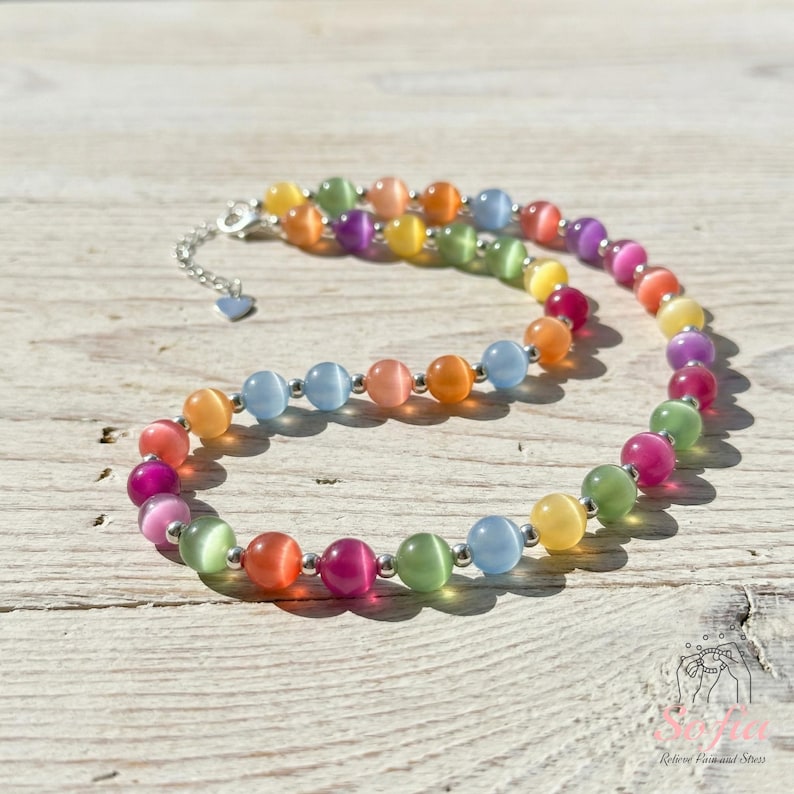 May include: A necklace featuring a strand of multicolored cat's eye beads in shades of pink, orange, yellow, green, and blue. The beads are separated by small silver-toned metal spacers, and the necklace includes a silver-toned metal chain extender.