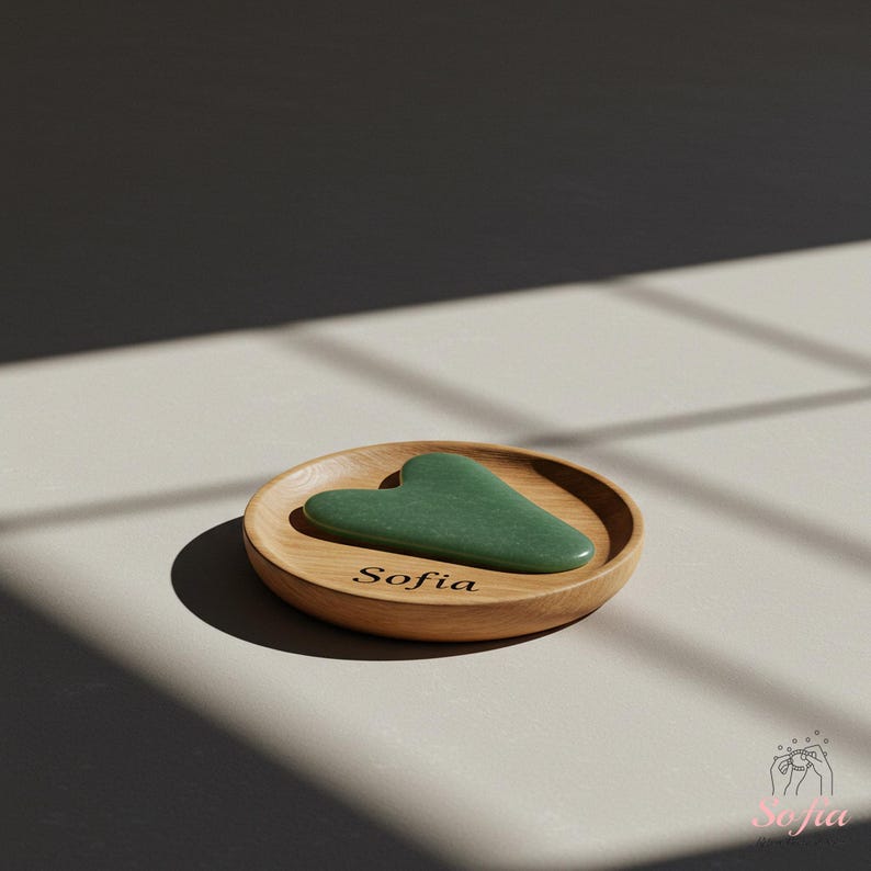 May include: A heart-shaped green jade gua sha tool sits on a small, round wooden dish. The dish is engraved with the name "Sofia". The background is a neutral colour with shadows.