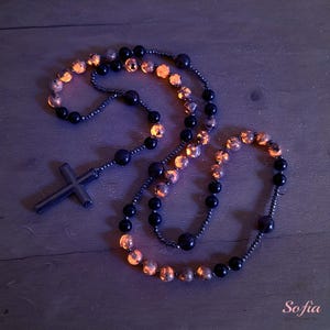 May include: A rosary with a black cross pendant and black beads. The rosary also features amber-colored beads. The beads are strung together with a dark-colored chain. The rosary is displayed on a dark wooden surface.