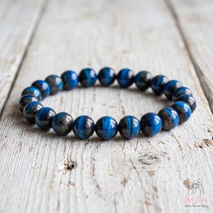 May include: A stretch bracelet composed of round, polished beads in shades of deep blue and charcoal gray. The beads exhibit a chatoyant, shimmering effect across their surfaces, arranged in a circular loop on a light-colored wooden surface.