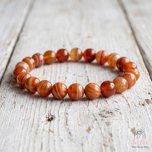 May include: A stretch bracelet composed of round, polished beads in varying shades of burnt orange, rust, and deep red. Each bead features distinct, concentric banded patterns in lighter and darker tones, creating a natural, layered appearance.
