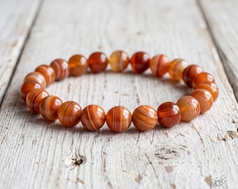 May include: A stretch bracelet composed of round, polished beads in varying shades of burnt orange, rust, and deep red. Each bead features distinct, concentric banded patterns in lighter and darker tones, creating a natural, layered appearance.